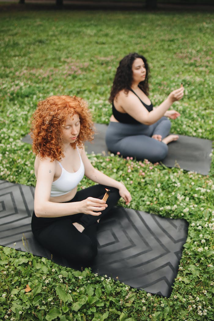 contact-02 Two women meditating on yoga mats in a park, embracing mindfulness outdoors.